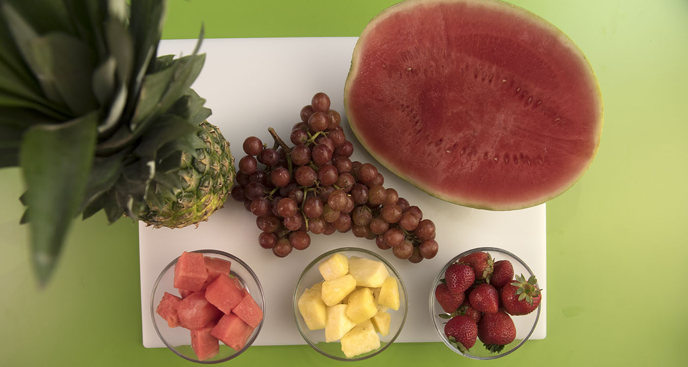 A sliced watermelon, a bunch of grapes, a whole pineapple and a bowl of cubed watermelon, a bowl of cubed pineapple, and a bowl of strawberries on a white cutting board.