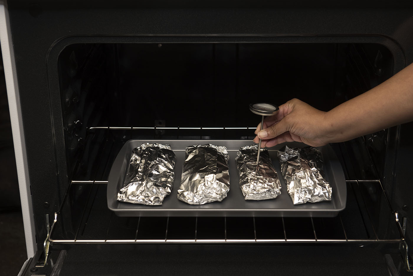 Four foil pouches on a baking sheet in an oven with a woman testing the temperature of one.