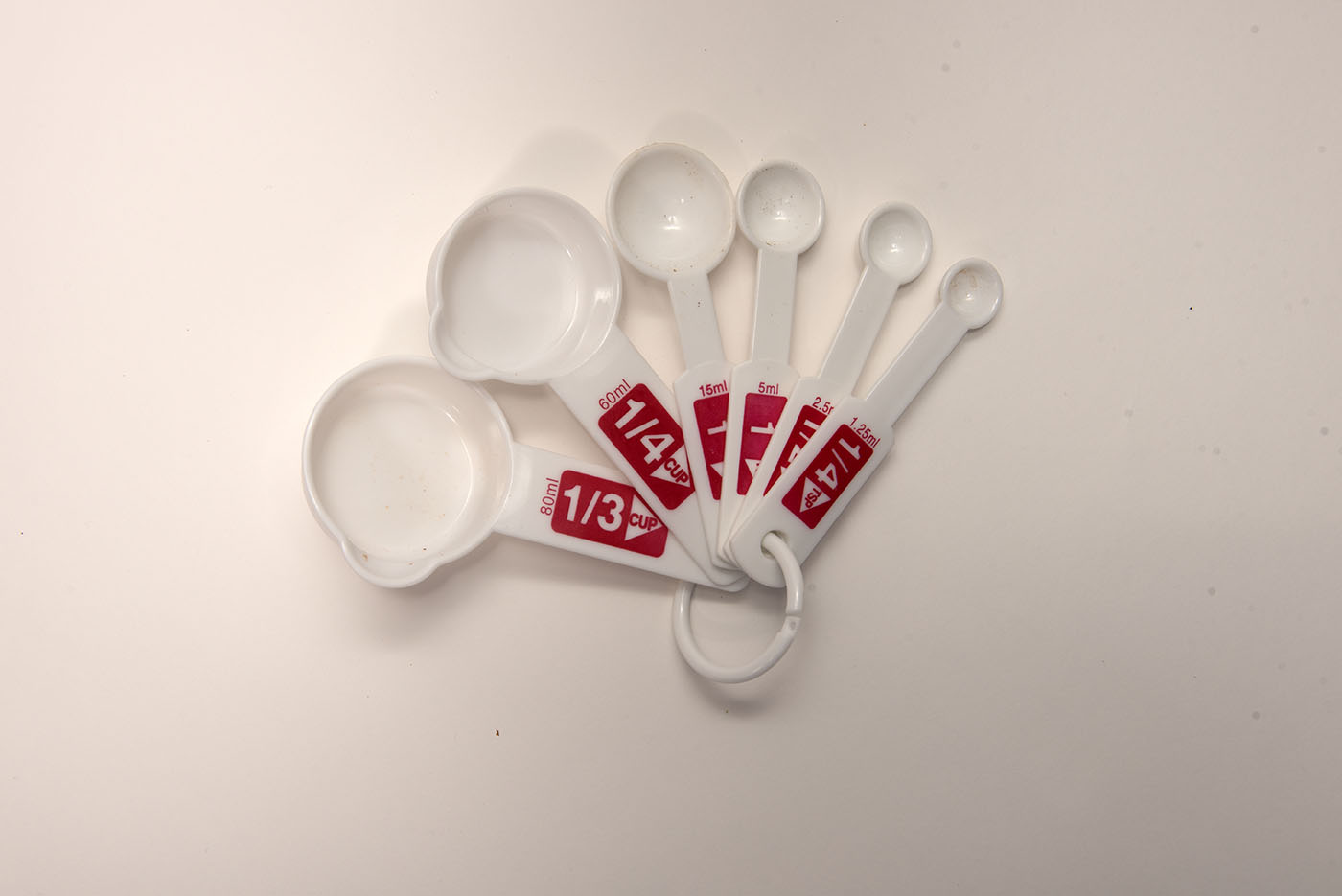 A set of white measuring cups with 1/3 cup, 1/4cup, 1 tablespoon, 1 teaspoon, 1/2 teaspoon, and 1/4 teaspoon written in red on the corresponding cup/spoon.