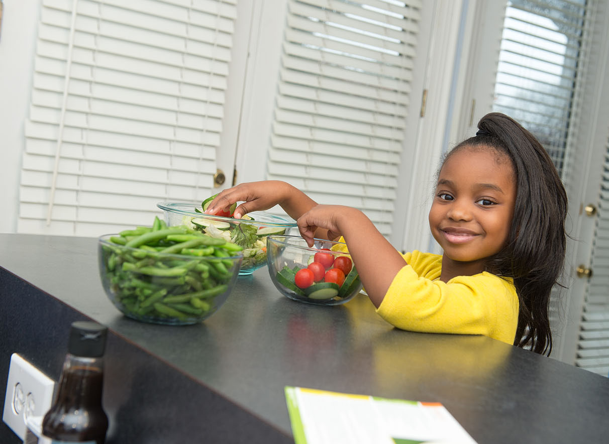 A smiling girl adds cherry tomatoes and sliced cucumbers to a larger bowl of salad greens. Snapped green beans are in a clear glass bowl are nearby.