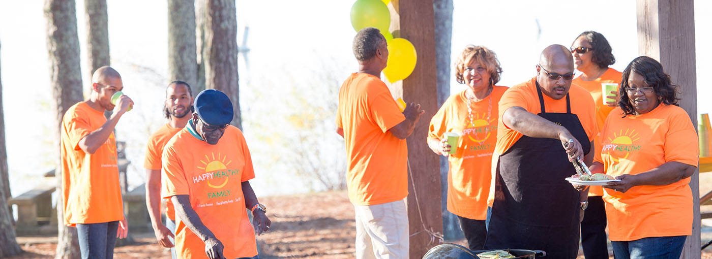 Six adults in orange shirts talking and grilling at a barbeque.