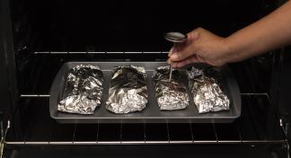 Hand with meat thermometer checking one of four foil-wrapped pieces of meat on baking sheet in oven