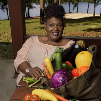 A woman smiles as she sits on a bench holding a large bag which contains squash, zuchinni, carrots, purple cabbage, egg plant, acorn squash, tomato, and greens.